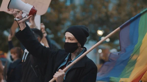 Mujer con máscara facial que habla en el megáfono mientras sostiene la bandera del arco iris. Demostración contra la dicriminación — Foto de Stock