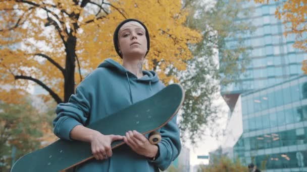 Мятежная молодая белая женщина со скейтбордом, кивая головой в знак одобрения в городском парке осенью — стоковое видео