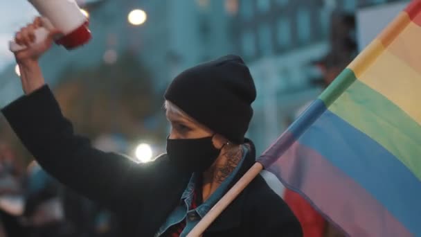 Жінка з маскою для обличчя, що тримає мегафон та веселковий прапор. Демонстрація проти дискримінації — стокове відео