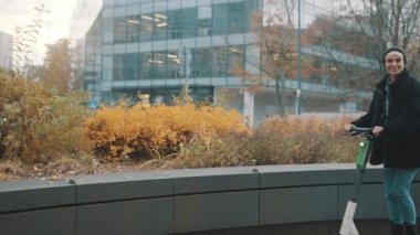 Kışın siyah ceketli, şapkalı ve maskeli genç bir kadın sonbaharda şehirde elektromanyetik scooter kullanıyor.
