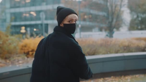 Mujer joven en chaqueta negra de invierno con sombrero y máscara facial montando scooter electico en el parque de la ciudad — Vídeo de stock