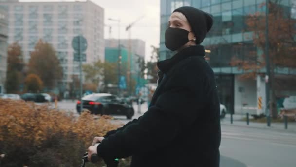Mujer joven en chaqueta de invierno negro con sombrero y mascarilla de montar scooter electico en la ciudad en otoño — Vídeo de stock