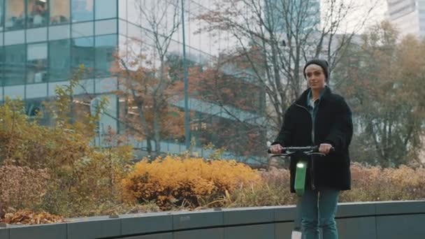 Kışın siyah ceketli, şapkalı ve maskeli genç bir kadın sonbaharda şehirde elektromanyetik scooter kullanıyor. — Stok video