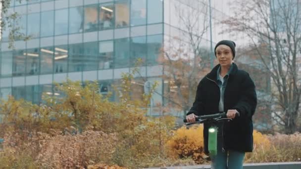 Şehir parkında şapkalı, siyah ceketli, elektromanyetik scooter kullanan genç bir kadın. Ekolojik toplu taşıma. Çevre hayatı kavramı, aktif yaşam tarzı — Stok video