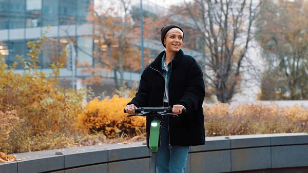 Mujer joven en chaqueta negra de invierno con sombrero de montar scooter electico en el parque de la ciudad. Transporte público ecológico. Concepto de vida ecológica, estilo de vida activo — Foto de Stock