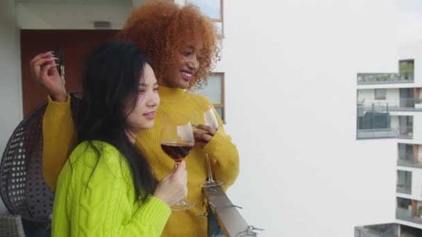 Un par de mujeres jóvenes interracial, mejores amigos o pareja lesbiana, bebiendo vino en el balcón. Africano americano mujer acariciando cabello de su asiático pareja — Vídeos de Stock