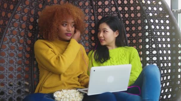 Νεαρές γυναίκες, καλύτερες φίλες μοιράζονται ακουστικά και ακούνε μουσική από το laptop, χαλαρώνουν στην κούνια στο μπαλκόνι με ποπ κορν — Αρχείο Βίντεο