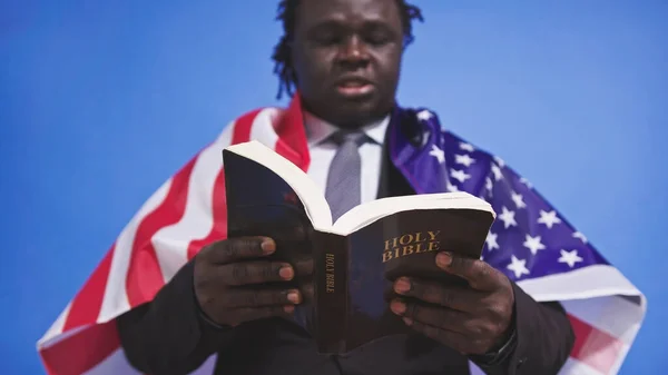 Close up, bíblia sagrada nas mãos do homem negro afro-americano em terno elegante com bandeira dos EUA sobre os ombros — Fotografia de Stock