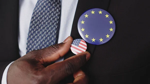 Close Up of Black Male Hand compare American avec l'insigne du drapeau de l'Union européenne sur un costume élégant. Economie et politique Concept, détail — Photo