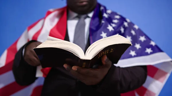 Крупный план, священная библия в руках черного африканского американца в элегантном костюме с флагом США на плечах — стоковое фото