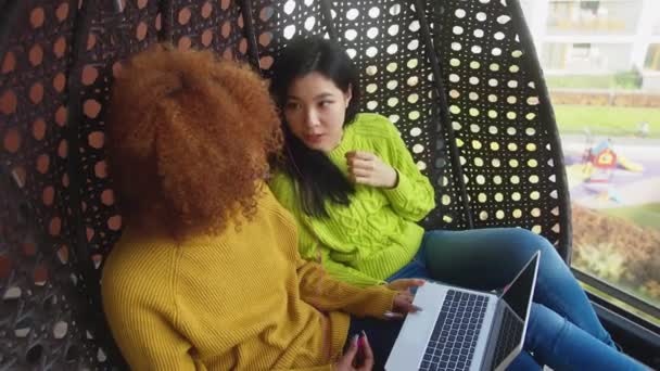 L'amitié multiraciale. Jeunes femmes africaines américaines et asiatiques bavardant sur la balançoire sur la terrasse annonce vérifier les médias sociaux sur l'ordinateur portable — Video