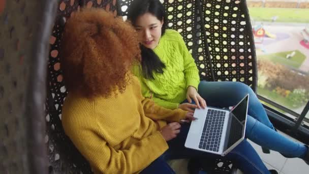 Amistad multirracial. Jóvenes afroamericanos americanos y asiáticos cotilleando en el columpio en la terraza y navegando por las redes sociales en el portátil — Vídeos de Stock