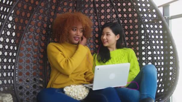 Jeunes femmes, nationalité différente, partage d'écouteurs et écoute de la musique depuis un ordinateur portable, détente au swing sur le balcon avec des popcorns — Video