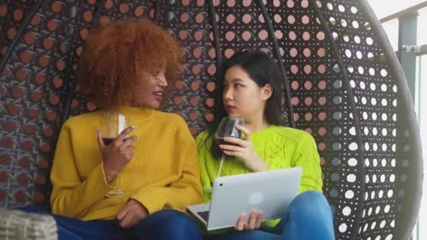 Två kvinnor slappnar av i gungan, bästa vänner dricker rödvin. Multietniskt förhållande eller homosexuellt förhållande — Stockvideo