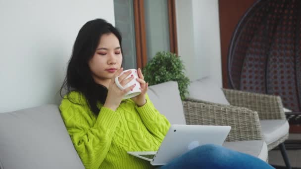 Concepto de relajación y bienestar. Mujer asiática joven bebiendo taza de té mientras descansa en el sofá del balcón con el ordenador portátil — Vídeo de stock