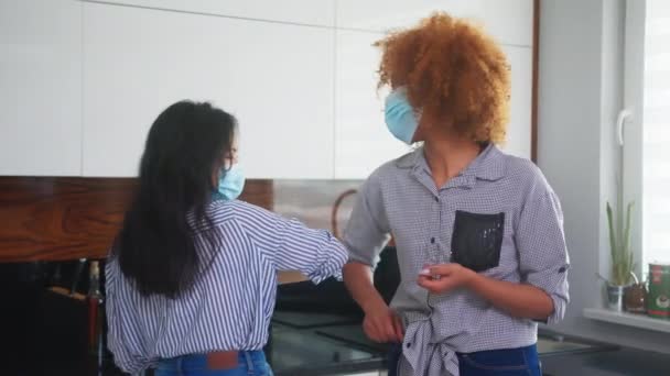 Ασιάτισσα και Αφρικανή Αμερικανίδα με ιατρικές μάσκες χαιρετιούνται μεταξύ τους με αγκώνες στην κουζίνα εργασίας στο διάλειμμα κατά τη διάρκεια του covid-19 — Αρχείο Βίντεο