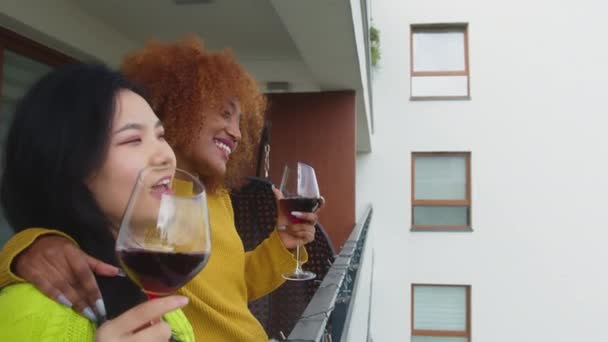 Dwie młode, międzyrasowe kobiety, najlepsze przyjaciółki lub lesbijki, pijące wino na balkonie, śpiewające i tańczące. Afroamerykanka głaszcze włosy swojego azjatyckiego partnera — Wideo stockowe