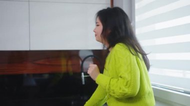 Genç Asyalı kadın mutfak dolabında oturmuş dans ediyor ve şarkı söylüyor.