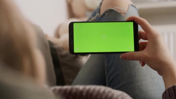 Jovem segurando smartphone com tela verde enquanto descansa no sofá e acenando. Chamada de vídeo — Vídeo de Stock