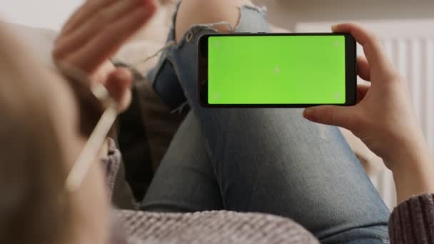Jovem segurando smartphone com tela verde enquanto descansa no sofá e acenando. Chamada de vídeo — Vídeo de Stock