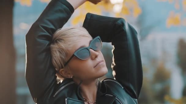 Ung kvinna med ljust hår och solglasögon poserar i parken med händerna över huvudet. Närbild — Stockvideo