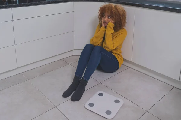 Devastated young african american black woman sitting on the flor with weight scale in front. Weight loss and mental health concept