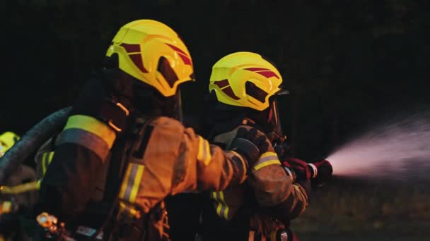 De cerca, dos bomberos apagan el fuego, rociando con manguera por la noche. — Vídeos de Stock