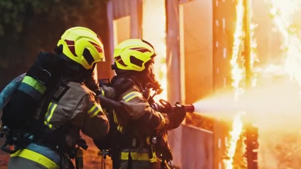 De cerca, dos bomberos apagan el fuego, rociando la casa con manguera por la noche. — Vídeos de Stock