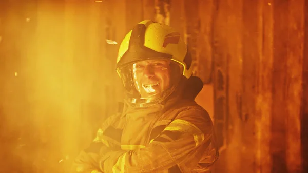Fiatal tűzoltó portréja az égő ház előtt, amint teljes egyenruhában mosolyog. — Stock Fotó