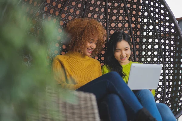 Dos mujeres relajándose en el columpio, y navegando por las redes sociales. Africano americano y asiático mujer, multirracial amistad concepto — Foto de Stock
