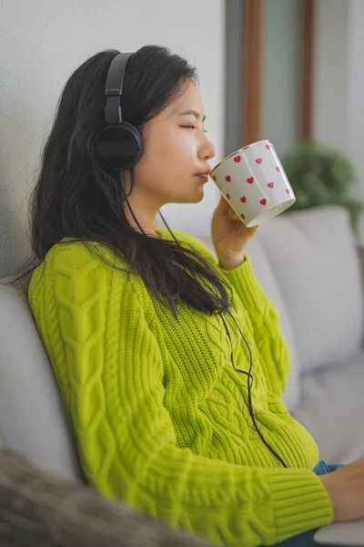 Concepto de relajación y bienestar. Mujer asiática joven bebiendo taza de té mientras descansa en el sofá del balcón con el ordenador portátil — Foto de Stock