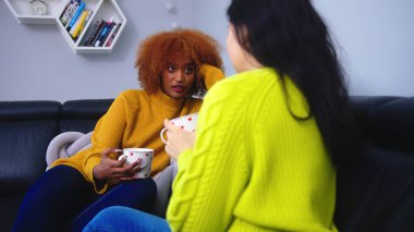 Kahve içen iki genç bayan arkadaş oturma odasında sohbet ediyorlar. Afrika kökenli Amerikalı ve Asyalı kadın oda arkadaşları dedikodu yapıyor. 