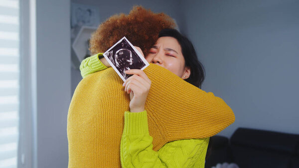 Многорасовая лесбийская пара ждет ребенка. Объятия с ультразвуковым снимком ребенка. Материнская поддержка