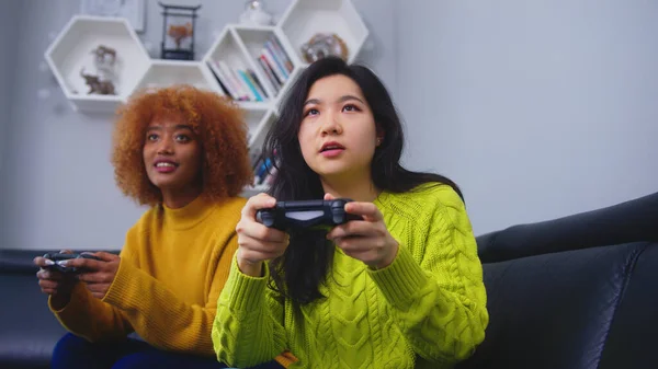 Lesbianas lgbt mujeres pareja jugar juegos en casa, Asiática y africana americana hembras tener divertido feliz momento juntos en sofá en sala de estar en la noche. — Foto de Stock