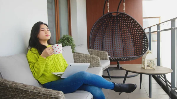 Jovem mulher asiática desfrutando de bela manhã em sua varanda com xícara de chá e laptop — Fotografia de Stock
