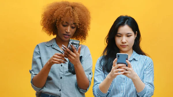 Dos mujeres jóvenes, afroamericanas negras y asiáticas usando teléfonos inteligentes — Foto de Stock