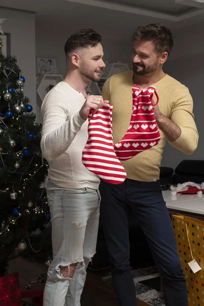 Χαρούμενο γκέι ζευγάρι ερωτευμένο να κρατάει χριστουγεννιάτικες κάλτσες και να αγκαλιάζεται μπροστά στο χριστουγεννιάτικο δέντρο. Παραμονή Χριστουγέννων και ομοφυλοφιλική σχέση — Φωτογραφία Αρχείου
