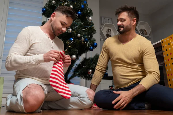 Παραμονή Χριστουγέννων. Ευτυχισμένοι γκέι άντρες που τσεκάρουν χριστουγεννιάτικες κάλτσες για δώρα. Καθισμένος κάτω από το χριστουγεννιάτικο δέντρο — Φωτογραφία Αρχείου