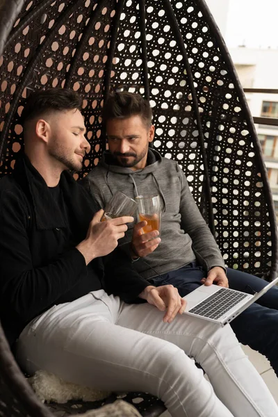 ¡Salud! Joven pareja gay guapo relajarse en el columpio en el balcón con cerveza y navegar por las redes sociales en el ordenador portátil —  Fotos de Stock