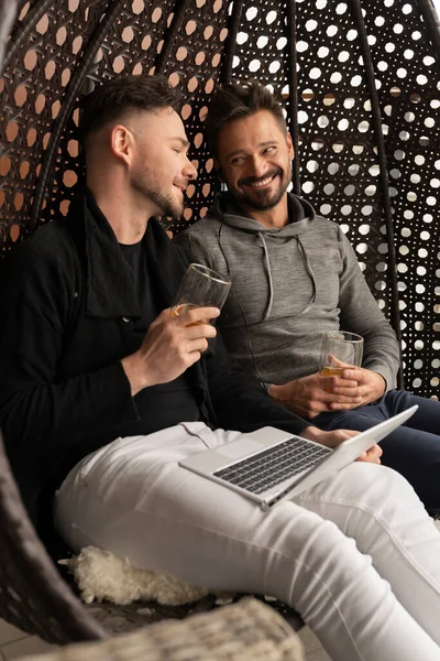 Joven pareja gay guapo riendo y relajándose en el columpio en el balcón con cerveza y portátil —  Fotos de Stock