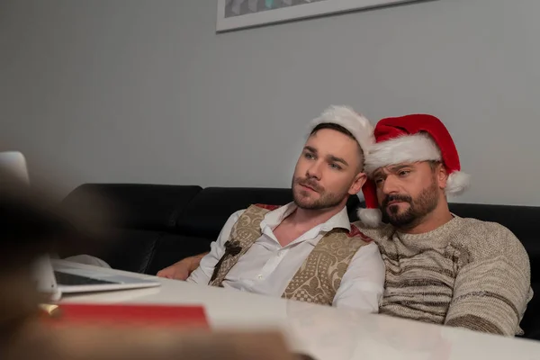 Молодая кавказская гей-пара в шляпах Санта-Клауса смотрит фильмы на ноутбуке в канун Рождества — стоковое фото