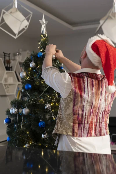 Πίσω όψη του νεαρού καυκάσιου με το καπέλο του Αϊ Βασίλη στολίζοντας το χριστουγεννιάτικο δέντρο — Φωτογραφία Αρχείου