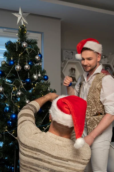 Atractiva pareja homosexual masculina que celebra el Año Nuevo, decorando juntos el árbol de Navidad. Familia feliz disfrutando de las vacaciones — Foto de Stock