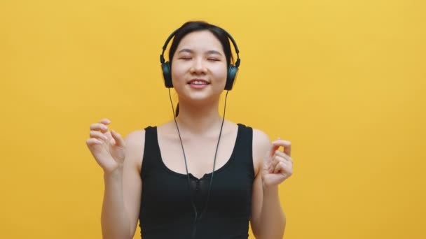 Счастливая молодая азиатка с наушниками, слушающая музыку и танцующая. Изолированный на желтом фоне — стоковое видео