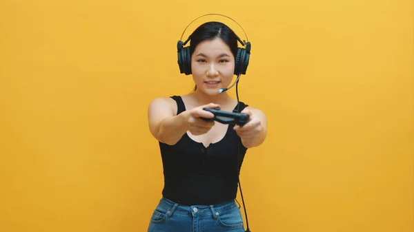 Enfocado mujer asiática joven con auriculares y controlador alegre jugando videojuegos. Aislado sobre fondo amarillo — Foto de Stock