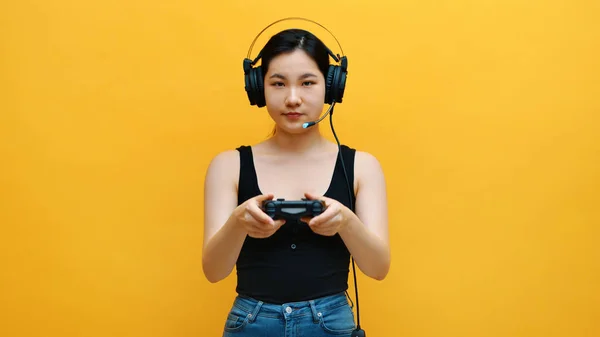 专注于年轻的亚洲女人与耳机和快乐控制器玩电子游戏。在黄色背景下隔离 — 图库照片