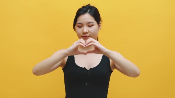 Feliz joven mujer asiática haciendo forma de corazón con sus manos. Aislado sobre fondo amarillo — Vídeo de stock