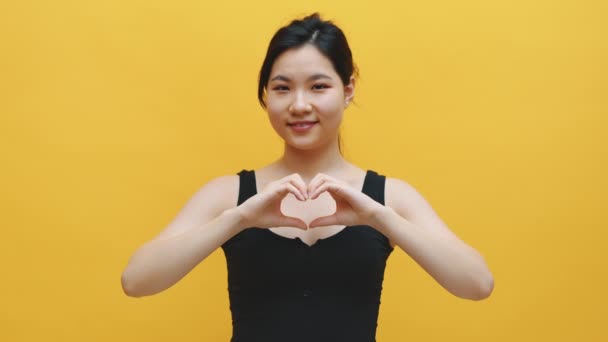 Linda jovem mulher asiática fazendo forma de coração com as mãos. Isolado sobre fundo amarelo — Vídeo de Stock