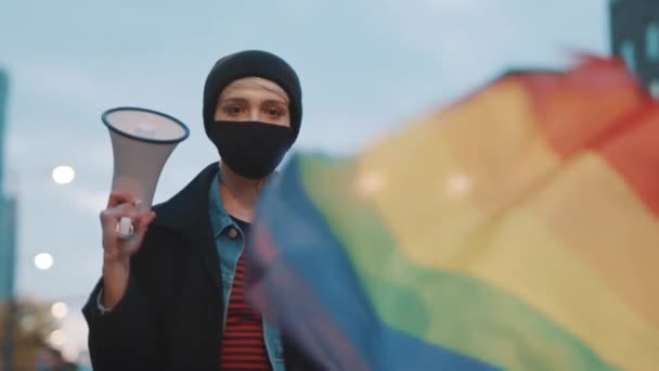 Жінка з маскою для обличчя, що тримає мегафон та веселковий прапор. Демонстрація проти дискримінації — стокове відео