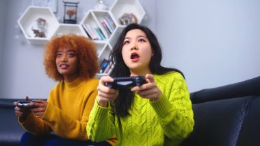 Evde video oyunu oynayan neşeli, mutlu, çekici kızlar. Odaklanmış Asyalı kadın Afrikalı Amerikalı siyahi arkadaşından oyunu kaybediyor.